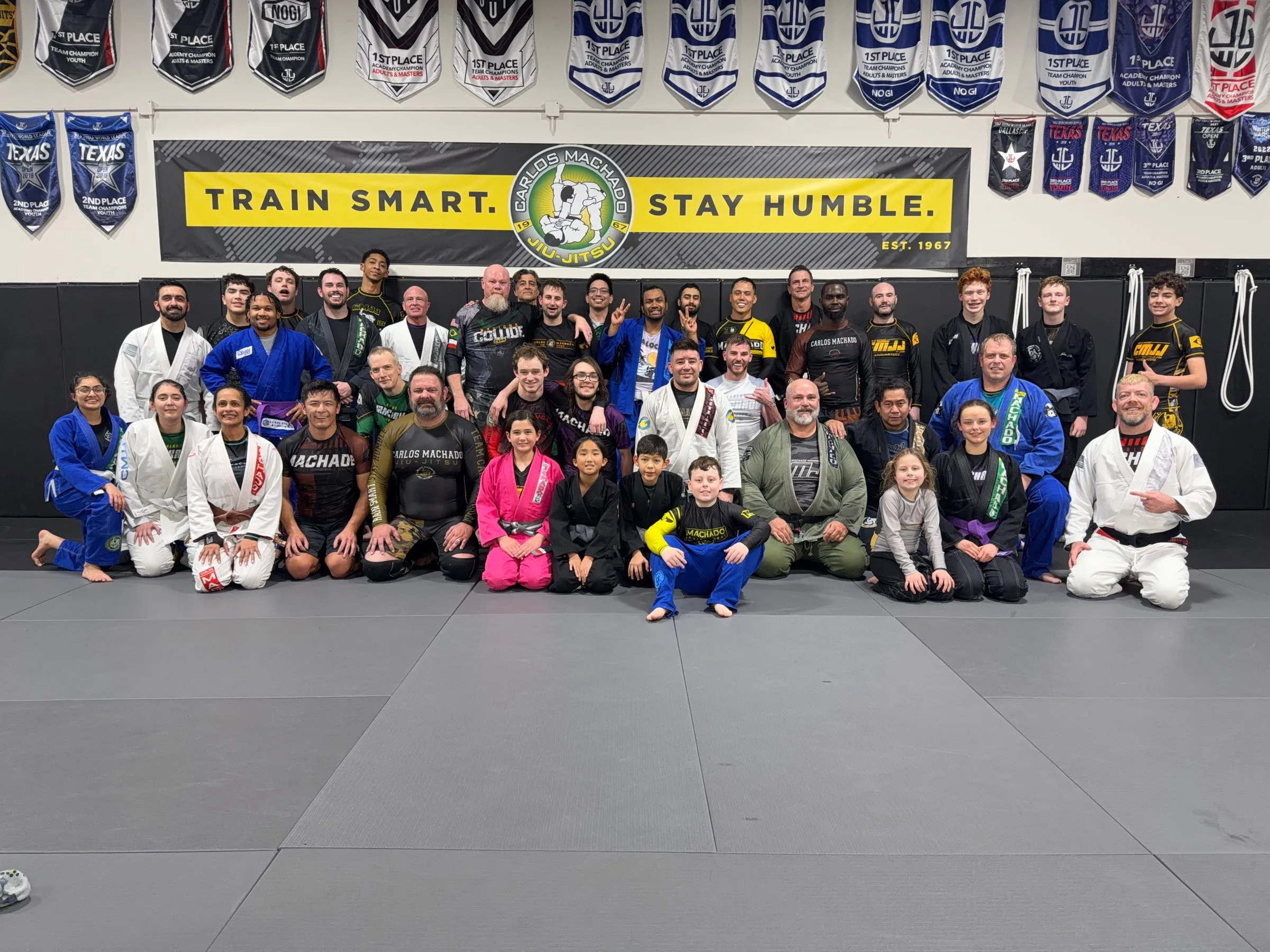 The whole Machado Method Carrollton school gathered on the mat under the Train Smart, Stay Humble banner