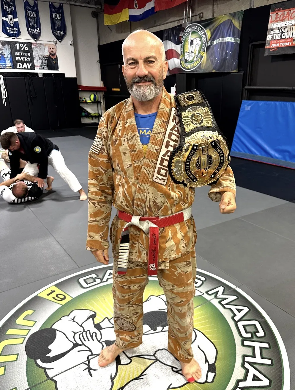 Master Carlos Machado in his gi holding a championship belt, standing on the Carlos Machado Jiu-Jitsu mat logo in Carrollton, TX