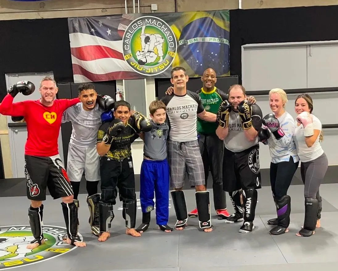 The striking class at Machado Method Carrollton standing together in boxing gloves and shin guards under the Carlos Machado banner